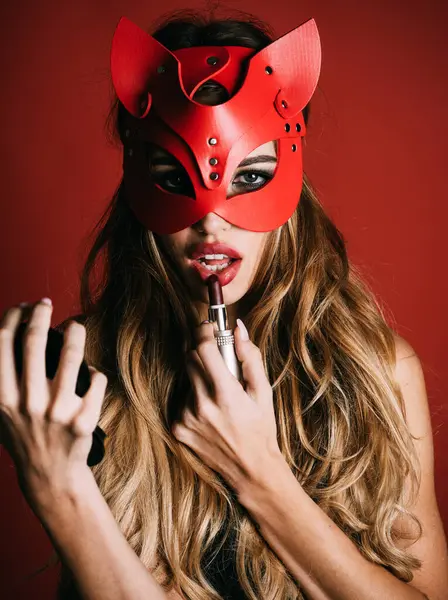 Cat woman. Sexy girl in cat mask with professional makeup preparing for party. Makeup for carnival, masquerade. Sensual woman applying dark red lipstick on lips holds mirror in hand. Closeup portrait
