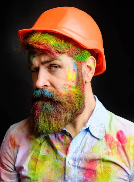 Closeup portrait of professional painter, decorator or builder in hard hat. Male painter in paint-splattered shirt. Handsome man with dyed hair and beard in protective helmet. Room painting job