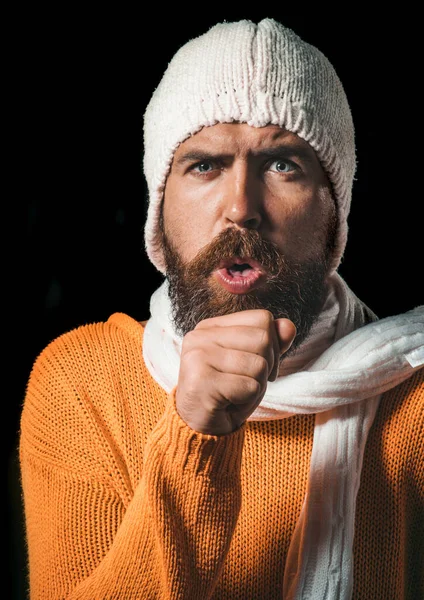 Bearded sick man in sweater, hat and scarf coughing. Handsome guy having sore throat and coughing into his fist. Unhealthy man wrapped in scarf have cough as symptom for influenza, cold or bronchitis