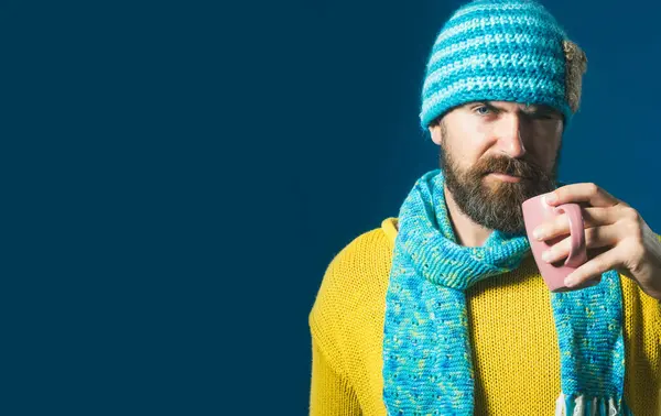 Serious stylish man in knitted sweater and hat wrapped in scarf with mug of coffee or tea in cafe. Handsome bearded man in fashionable winter clothes drinking hot beverage. Copy space for advertising