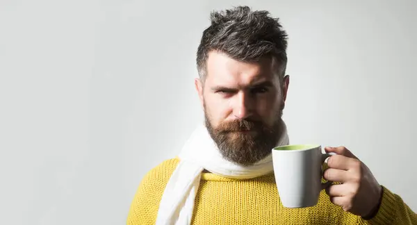 Serious man in knitted sweater wrapped in scarf with cup of coffee or tea. Autumn or winter time. Handsome bearded man in warm clothes drinking beverage in cafe or bistro. Copy space for advertising