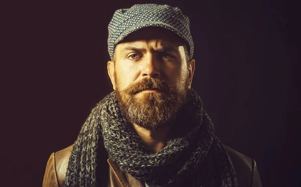 Fashion male model in winter outfit. Serious bearded man in leather jacket, gray cap and scarf. Fashion for men. Stylish handsome man with beard and mustache in autumn-winter clothes. Winter knitwear