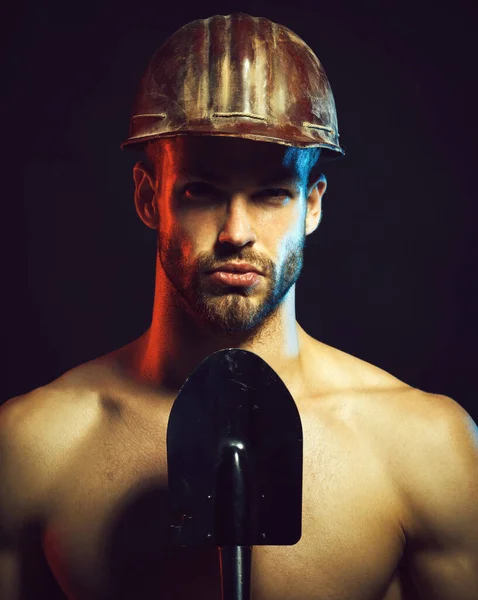 Sexy bearded builder, construction worker in protective hard hat with spade. Foreman, repairman or miner in protective helmet with shovel. Mechanical or industrial worker in safety hardhat with spade
