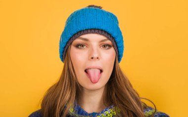 Örgü şapkalı ve atkılı güzel kızın dilini gösteren yakın plan portresi. Sonbahar-kış giysileri içinde moda kadın modelleri. Temiz tenli, mükemmel makyajlı, yün şapkalı ve atkılı genç bir kadın.