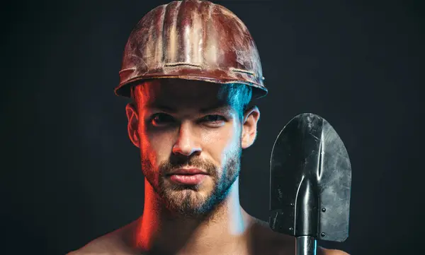 Closeup portrait of construction worker in safety hard hat with spade. Serious bearded man in construction helmet with shovel. Sexy handsome builder, miner, repairman in protective hardhat with spade
