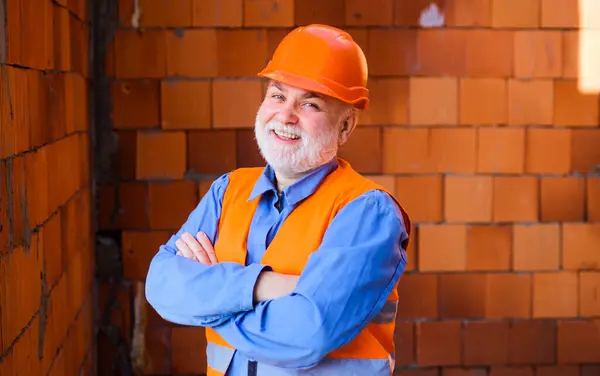 Building industry. Smiling builder man, engineer, architect in hard hat on construction site. House construction. Handsome bearded inspector, foreman, craftsman in protective helmet with crossed arms