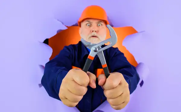 Professional plumber, construction worker or builder in hard hat with adjustable wrench looking through paper hole. Repairer in uniform with with adjustable spanner ready to work. Tools for repair