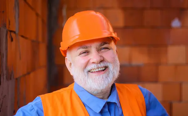Closeup portrait smiling construction worker in hard hat. Bearded workman in protective helmet at building site. Male architect, contractor or builder in safety hardhat working on house construction