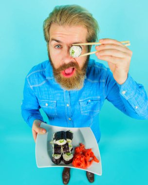 Sushi teslimatı. Sıradan kot gömlekli, ciddi sakallı bir adam gözlerini suşi rulosuyla kapatıyor. Geleneksel Japon yemekleri. Bir tabak lezzetli maki suşi ve turşulu zencefilli yakışıklı erkek. Deniz yiyeceği