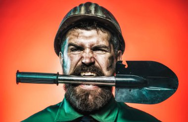 Madencilik, ağır sanayi ve inşaat konsepti. Endüstriyel işçi, dişlerinde kürekle kasklı bir madenci. Kızgın sakallı adam işçi üniforması ve miğferi içinde istihkamcı küreği ya da askeri küreği olan.