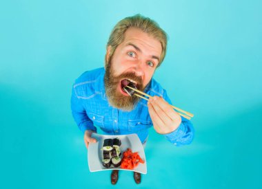 Sıradan kot gömlekli sakallı bir adam yemek çubuklarıyla suşi yiyor. Sushi teslimatı. Geleneksel Japon yemekleri. Bir tabak lezzetli maki suşi ve turşulu zencefilli yakışıklı erkek. Deniz yiyeceği