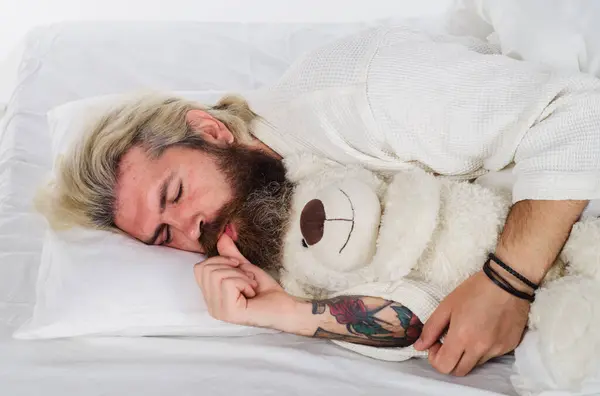 Bearded man sleeping with plush teddy bear relaxing in bed. Good night. Sleepy man in pajamas sleeping in bed at home with teddy bear toy. Handsome guy sleeping at bedroom and hugging soft teddybear