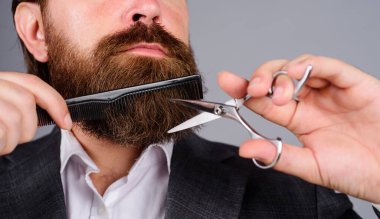 Berber dükkanı prosedürleri. Profesyonel sakal bakımı. Takım elbiseli şık sakallı adam sakalını ve bıyığını makas ve tarakla kesiyor. Erkekler için bir salon. Berber aletleriyle kuaför. Berber dükkanı reklamı.