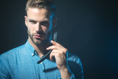 Berber dükkanı aksesuarları. Düz usturalı kot gömlekli ciddi sakallı bir adam. Profesyonel sakal bakımı. Sakallı ve bıyıklı şık bir adam ustura tutuyor. Berber dükkanı reklamı için yer kopyala