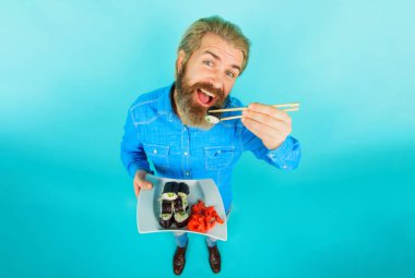 Sakallı adam bambu çubuklarıyla suşi yiyor. Bir tabak suşi rulosu ve turşulu zencefilli kot gömlekli yakışıklı bir erkek. Sushi teslimatı. Gülümseyen adam geleneksel Japon yemeklerini seviyor.