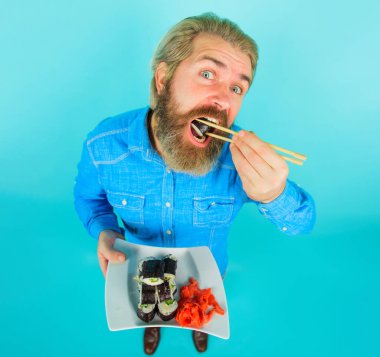 Sakallı adam lezzetli suşi rulolarını çubuklarla yer. Suşi menüsü. Lezzetli maki suşi rulosu ve tabakta zencefil turşusu olan kot gömlekli yakışıklı bir adam. Geleneksel Japon yemekleri. Suşi teslimatı