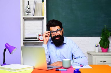 Sınıftaki gülümseyen erkek öğretmen dizüstü bilgisayarla çalışıyor. Gözlüklü üniversite öğrencisi internette ders çalışıyor. Kolejde ya da lisede e-öğrenim. Çevrimiçi test. Eğitim ve bilgi