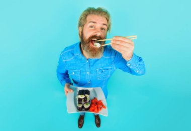 Geleneksel Japon mutfağı. Kot gömlekli gülümseyen sakallı adam yemek çubuklarıyla lezzetli suşi ruloları yiyor. Lezzetli maki suşi ruloları ve turşulu zencefilli turşulu yakışıklı bir erkek. Suşi teslimatı