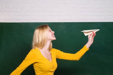 Kız öğrenci origami uçağıyla karatahtanın yanında oynuyor. Seyahat hayalleri. Gülümseyen kız kağıt uçak fırlatıyor. Genç bir kadın sınıfta kağıt uçak fırlatıyor. Mutlu kız kağıt uçakla eğleniyor.
