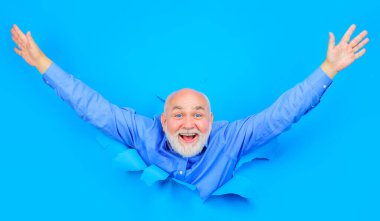 Mutlu sakallı adam mavi kağıttaki delikten bakıyor. Ellerini kaldırmış, şık gömlekli, gülümseyen yaşlı adam. Kırma kağıdı. Metin veya slogan için boşluk kopyala. İndirim, sezon indirimi. Kara Cuma. Reklamcılık