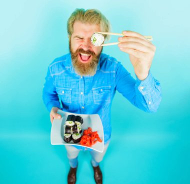 Sakallı adam lezzetli yemek çubuklarıyla suşi yiyor. Geleneksel Japon mutfağı. Lezzetli maki suşili rulo ve turşulu zencefilli yakışıklı erkek. Sushi teslimatı. Japon yemeği. Seçici odak