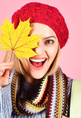 Gözlerini sarı akçaağaç yaprağıyla kaplayan gülümseyen kadının yakın plan portresi. Sonbahar modası. Yünlü şapka, atkı ve sonbahar yapraklı süveter giyen mutlu kız. Şık ve sıcak bir sonbahar modeli. Sonbahar indirimi
