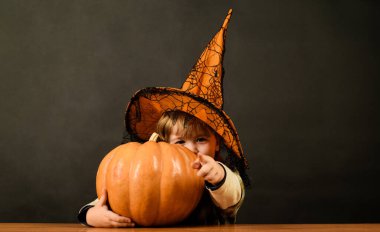 Cadılar Bayramı şapkalı Cadılar Bayramı çocuğu ve balkabağı seni işaret ediyor. Cadılar Bayramı kostümü içinde balkabaklı şirin bir çocuk. Gülümseyen küçük çocuk Cadılar Bayramı partisine hazırlanıyor. Sezon satışları