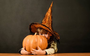 Cadılar Bayramı çocuğu cadı şapkasıyla koca balkabağına sarılıyor. Cadılar Bayramı için hazırlık. Cadılar Bayramı karnavalı kostümü içinde balkabaklı şirin bir çocuk. Cadılar Bayramı indirimi, alışveriş ve indirim.