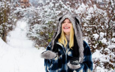 Ekose ceketli, kürklü ve eldivenli gülümseyen kadın kış parkında yürüyor. Soğuk kış havası. Kar yağışlı kış gününde, kalın giysiler içinde güzel bir Noel kadını. Kış ormanlarında örnek bir kız.
