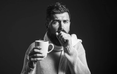 Sakallı adamın boğazı ağrıyor, yumruğuna öksürüyor ve fincandan iyileştirici içecek içiyor. Hasta adam öksürüyor. Süveter ve eşarplı hasta bir adam ve bir fincan sıcak tıbbi öksürük çayı. Siyah ve beyaz