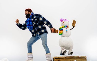 Noel ve yeni yıl kutlaması. Ekose paltolu mutlu sakallı adam ve buz patenli kardan adam. Aktif eğlence, kış sporu. Kışlık elbiseli bir adam şapkalı, atkılı ve eldivenli komik bir kardanadamla koşuyor.