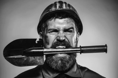 Dişinde kürek olan koruyucu kasklı bir erkek müteahhit ya da madenci. Madencilik, ağır sanayi, inşaat konsepti. Üniformalı kızgın sakallı bir adam portresi ve kasketli bir kürek. Siyah ve beyaz