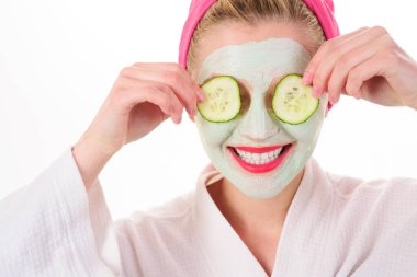 Kil kozmetik maskeli ve gözlerinde salatalık dilimleri olan gülümseyen bir kadın. Güzellik ve cilt bakımı evde. Cilt maskesi ve gözlerinde salatalık olan genç bir kız. Spa tedavileri ve sağlık