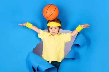 Kafasında top olan gülümseyen çocuk mavi kağıttaki delikten bakıyor. Spor malzemeleri. Basketbol toplu sarı tişörtlü küçük basketbolcu. Basketbol antrenmanı. Basketbol oynayan sevimli çocuk.
