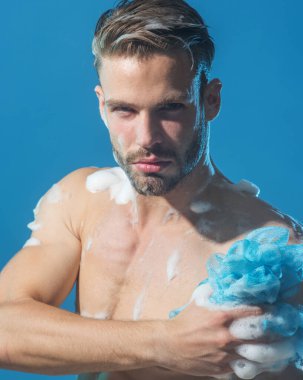 Yakışıklı, ciddi bir adam vücudunu sabunlu sünger ve nemlendirici jelle yıkıyor. Vücut bakımı için erkek kozmetiği. Sabah rutini. Cilt bakımı ve vücut bakımı. Banyoda duş alan kaslı, tıraşsız adam.