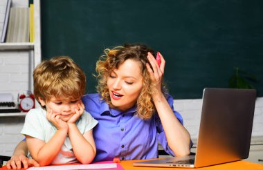 Bayan ilkokul öğretmeni küçük okul çocuğuna ev ödevinde yardım ediyor. Bilgi, eğitim ve öğrenme. İlkokul öğrencisi ve sınıfta özel öğretmen. Anne okulunda birlikte okuyan bir öğrenci.