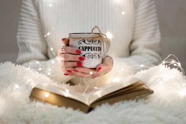 Flowers, old books and garlands. Winter calm. The smell of old books. Light, warm and cozy card. A girl holding a cup of hot cappuccino in bed.