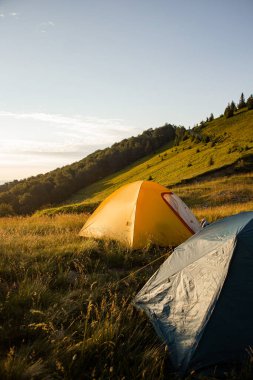 Gün batımında manzaralı bir dağ yamacına kurulmuş iki renkli çadır. Çevresi yeşil çimler, orman ve yürüyüş patikasıyla çevrili, doğada huzurlu bir yaz kampı. Açık hava macerası ve özgürlük.