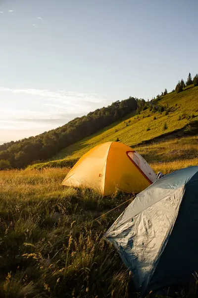 Gün batımında manzaralı bir dağ yamacına kurulmuş iki renkli çadır. Çevresi yeşil çimler, orman ve yürüyüş patikasıyla çevrili, doğada huzurlu bir yaz kampı. Açık hava macerası ve özgürlük.