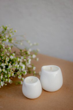 Minimalist beyaz seramik mumlar ve kahverengi arka planda küçük beyaz çiçekler. Sıcak ve sakin bir atmosfer, doğal ışık, yumuşak odaklanma. Ev dekorasyonu ve aromaterapi konsepti.