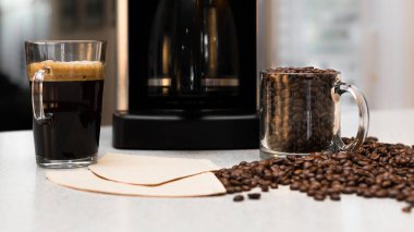 Cup of fresh coffee on kitchen counter table and blurred background. Pile of coffee beans, freshly made pot, and mug close up. Coffee cup filled with coffee beans