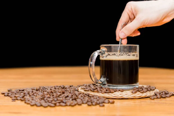 Man stirring a cup of coffee with a spoon. Coffee mug on wooden table and pile of coffee beans isolated on black background with copy space for text