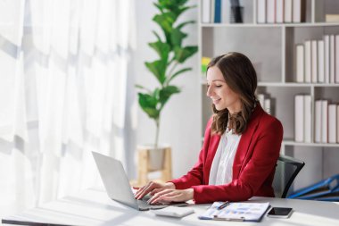Concept of business working, Businesswoman wearing red suite smiling while using computer laptop for checking business analytic document  report  in her workstation.