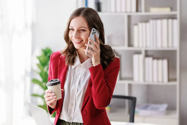 Concept of business working, Businesswoman wearing red suite standing while using smartphone talk with her client  about business analytic report and holding coffee cup in her hand.