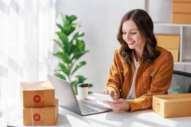 Paket teslimatı ve çevrimiçi satış kavramı, kadın satıcı veya perakendecinin günün satışlarını kontrol etmek için online mağaza kullanması ve paketlenip müşteriye teslim edilmeye hazır olması.
