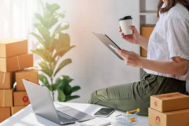 Paket teslimatı ve çevrimiçi satış kavramı, Retailer, bir çevrimiçi mağazada günün satışlarını kontrol etmek için bilgisayar tableti kullanırken, bir yandan da müşteriye göndermeye hazırlanan paket kutulu kahve fincanı bulunduruyor.