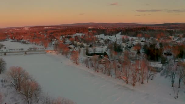 氷結した雪に覆われた川沿いの風光明媚な小さな町の冬の空中風景日没 — ストック動画