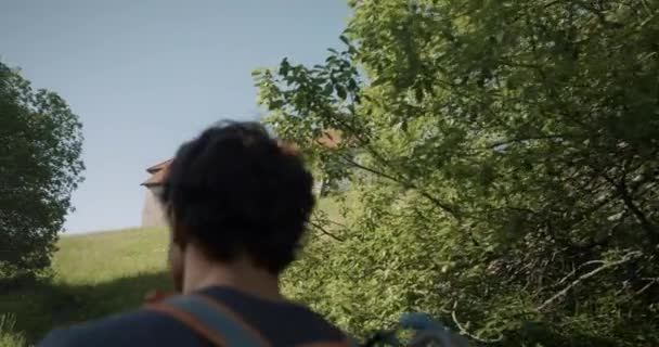 男ハイカーは丘の上にバックパックで歩くカム 石で作られた古い教会や塔を見て スロベニアで彼の前に青空と草や木の間で サイドショットを追跡 — ストック動画
