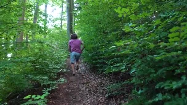 赤いTシャツを着た若い男がゆっくりと森の中を小道を走っている カメラは彼の後を追っています — ストック動画