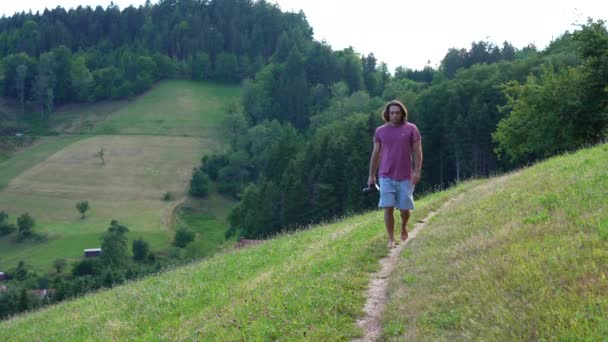 赤いTシャツと長い髪の若い男は 晴れた夏の日に丘の上の小さなパス上のカメラに向かってスローモーションで裸足で歩く 背景にある山と森 — ストック動画